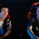 Two young people wearing headsets play games in illuminated gaming chairs