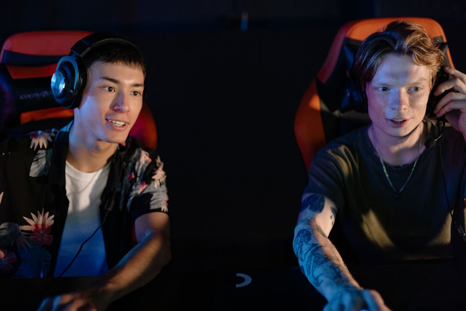 Two young people wearing headsets play games in illuminated gaming chairs